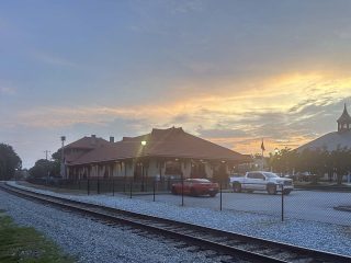 GVL A historic train station with a red roof stands next to railroad tracks at sunset. Two vehicles are parked in front, and the sky glows softly, as a fence marks the boundary—reflecting recent City Council discussions on the Greer budget.