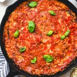 GVL A cast iron skillet filled with Grilled Eggplant Parmesan, topped with tomato sauce and fresh basil leaves, set on a light surface next to a striped towel.