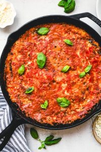 GVL A cast iron skillet filled with Grilled Eggplant Parmesan, topped with tomato sauce and fresh basil leaves, set on a light surface next to a striped towel.
