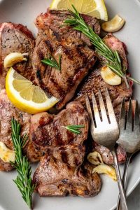 GVL Grilled Lamb Chops garnished with fresh rosemary, lemon wedges, and roasted garlic cloves on a plate. Two metal forks rest beside this delicious Rosemary Lamb Chops recipe.