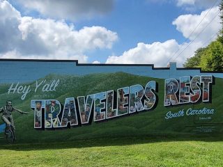 GVL A mural on a wall reads Hey Yall Welcome to Travelers Rest, South Carolina against a backdrop of green hills and blue sky with clouds. The letters, filled with illustrated scenes from Travelers Rest, celebrate the town’s community spirit.