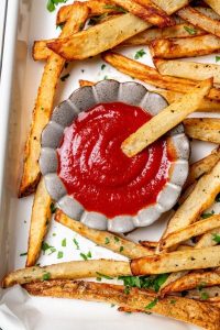 GVL A small gray bowl filled with homemade ketchup sits on a tray surrounded by crispy golden French fries. One fry is dipped into the ketchup, and chopped green herbs are sprinkled around.