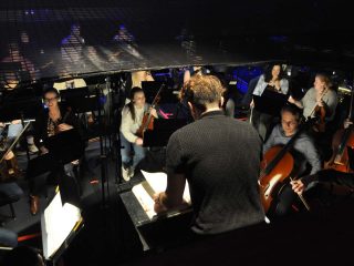 GVL A conductor leads the Greenville Symphony in a dimly lit orchestra pit, with musicians playing various string instruments and reading sheet music during this classical concert.