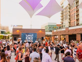 GVL A large crowd of people dances outdoors in downtown Greenville’s lively urban plaza for Salsa at Sunset on June 27, with geometric fabric canopies overhead and a band performing on a small stage.