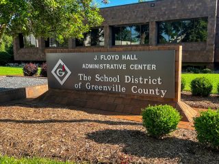 GVL A sign in front of a building reads J. Floyd Hall Administrative Center, Greenville County Schools Board of Trustees, The School District of Greenville County, surrounded by landscaping and greenery.