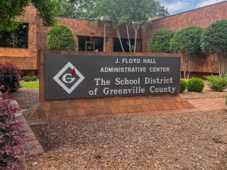 GVL A large sign outside a brick building reads “J. Floyd Hall Administrative Center, Greenville County Schools,” surrounded by neatly trimmed bushes and landscaping, highlighting the administrative hub for district budget decisions.