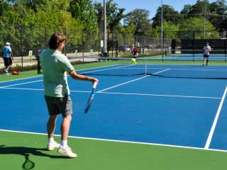 GVL A man in a light green shirt and shorts prepares to hit a tennis ball with his racket on the recreational courts at Cleveland Park in Greenville, while another player stands on the opposite side.