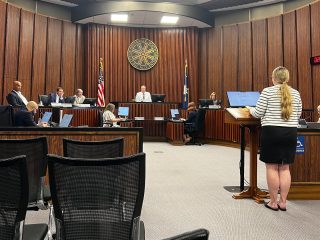 GVL A woman stands at a podium addressing the City Council in a wood-paneled chamber, discussing Greenville High’s parking expansion, with American flags and empty chairs visible in the foreground.