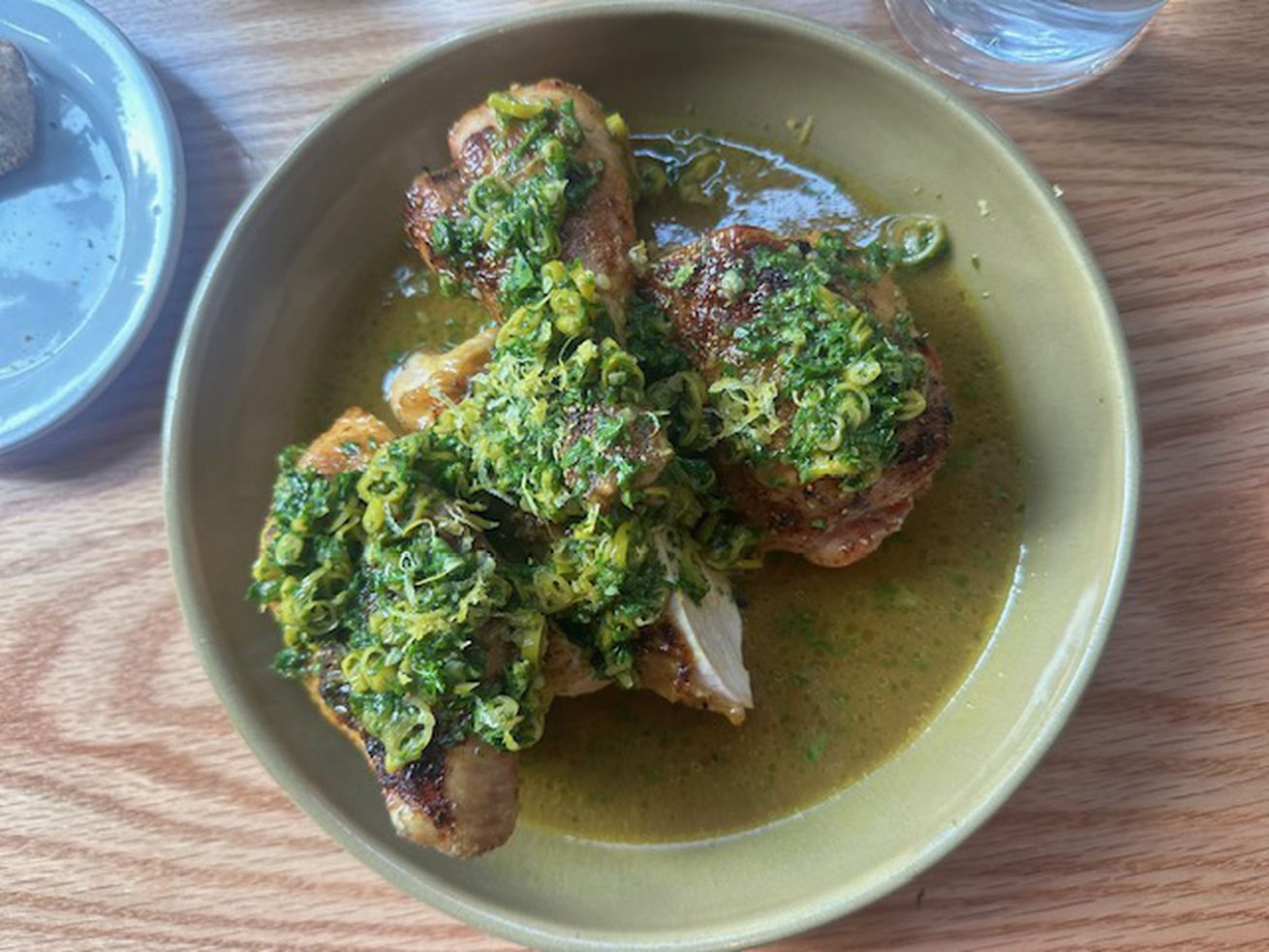 GVL A bowl of cooked chicken pieces topped with a green herb sauce, served in a light brown bowl on a wooden table—one of NY’s Best Dishes according to Editors. A glass of water and a plate with bread are partially visible nearby.