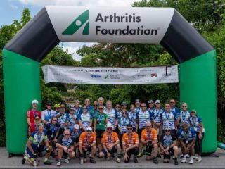 GVL A group of cyclists in matching jerseys poses under a large inflatable arch with the Arthritis Foundation logo at the Carolina Hills Classic cycling event, outdoors with trees in the background. Some wear blue jerseys and others wear orange.