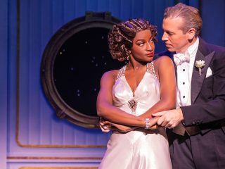 GVL A woman in a glamorous white gown stands with her arms crossed as a man in a tuxedo gently holds her from behind. They are on stage, in front of a nautical-themed backdrop evoking the Broadway style of Some Like It Hot.