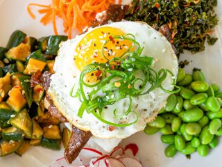 GVL A colorful plate of food from STIR, featuring a sunny-side-up egg topped with scallions and sesame seeds, surrounded by edamame, cucumber, carrot, kale, and sliced radishes—perfect for a vibrant downtown Father’s Day brunch.