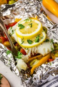 GVL A Mediterranean flavor shines in these fish packets: baked white fish topped with lemon slices and fresh herbs, nestled among bell peppers and cherry tomatoes. A fork rests beside the vibrant meal inside the packet.