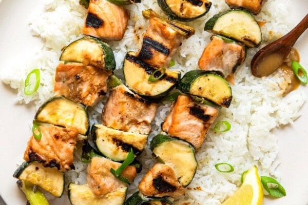 GVL Miso Salmon Skewers and zucchini served on a bed of white rice, garnished with sliced scallions and a lemon wedge, with a small bowl of sauce on the side.