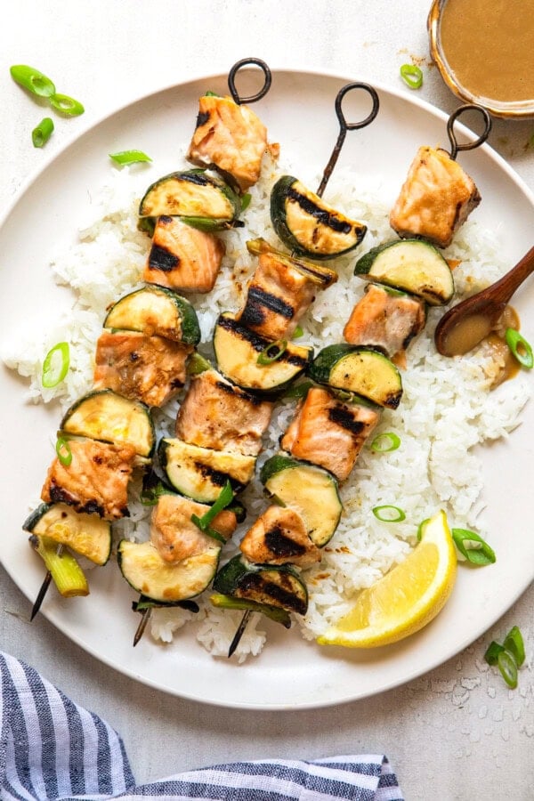 GVL Miso Salmon Skewers and zucchini served on a bed of white rice, garnished with sliced scallions and a lemon wedge, with a small bowl of sauce on the side.