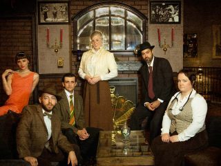 GVL Six people in vintage clothing pose in a dimly lit, elegant room with a fireplace and art deco details. With serious expressions, they evoke Centre Stage in a 1920s setting—where murder, plain and simple, might be at the heart of their gathering.
