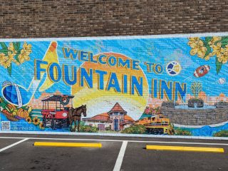 GVL Colorful mural on a brick wall in Fountain Inn reads “Welcome to Fountain Inn,” featuring a horse-drawn carriage, train, fountain, flowers, football, paintbrush, and bright sun against a blue sky—a vibrant nod supported by the City Council.