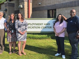 GVL Five adults stand smiling in front of a sign that reads Employment and Education Center at the Phoenix Center. They are outdoors on a sunny day, with a brick building and grass visible in the background, ready to explore new careers.