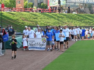GVL A group of people in matching shirts walk along a baseball field holding a Share the Magic banner during TD Synnex’s Day at the Drive community event. Spectators and greenery are visible in the background.
