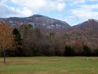 GVL A grassy field with scattered trees in the foreground and forested hills leading up to a tall, rocky mountain ridge under a partly cloudy sky, showcasing the beauty of land preservation and conservation in action.