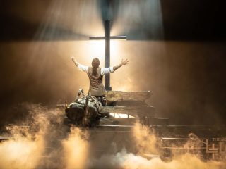GVL A person with outstretched arms kneels on a stage in front of a large cross, bathed in dramatic light and swirling fog, capturing the powerful, emotional atmosphere of a Logos Theatre production of Pilgrim’s Progress.