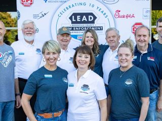 GVL A group of men and women smile in front of a banner celebrating 10 years of Law Enforcement Appreciation Day, wearing event shirts with sponsor logos visible in the background.