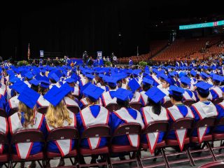 GVL Rows of Riverside Class of 2025 graduates in blue caps and gowns sit facing a stage during the graduation ceremony in a large indoor arena, with a crowd in the background capturing photos.