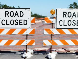 GVL Two Road Closed signs with orange and white striped barricades block Andrews Street. An orange warning light and plastic mesh fencing indicate sewer main work and lane closures ahead.