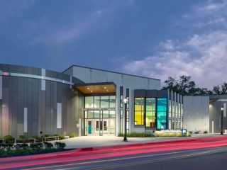 GVL Modern building with large glass windows and vertical panels, illuminated at dusk—home to the SC Children’s Theatre. Red and white light trails from passing cars streak past, while trees and a cloudy sky frame this artistic director’s vision.