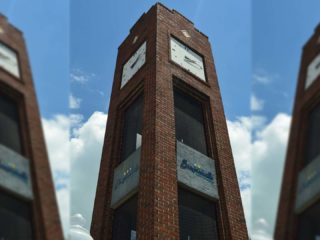 GVL A tall, brick clock tower in Simpsonville stands against a partly cloudy sky. The clock face is visible near the top, with signs—including recent short-term rental ordinances—mounted on the tower. The photo is taken from a low angle.