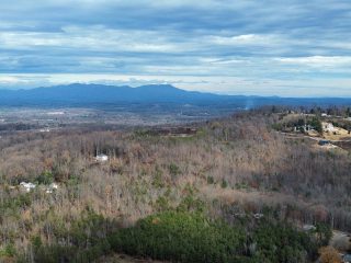 GVL Aerial view of a hilly landscape near Paris Mountain with scattered houses, leafless trees, and evergreens under a cloudy sky, reflecting the impact of zoning restrictions set by Greenville County Council, with distant mountains on the horizon.