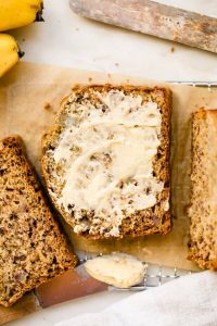 GVL A slice of banana bread with butter spread on top sits on parchment paper next to other slices. A butter knife and some bananas are visible in the background, perfect for enjoying this sourdough discard banana bread treat.