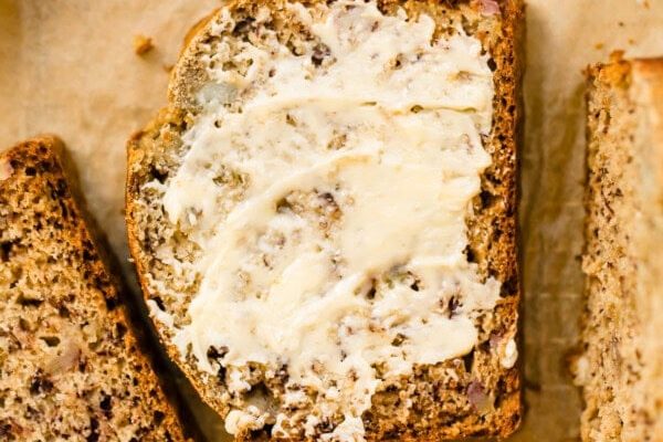 This Sourdough Discard Banana Bread Is So Good, I Make It Every Week! GVL A slice of banana bread with butter spread on top sits on parchment paper next to other slices. A butter knife and some bananas are visible in the background, perfect for enjoying this sourdough discard banana bread treat.