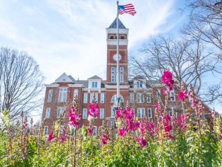 GVL A historic brick building with a clock tower stands behind blooming pink flowers and a flagpole with the American flag at Clemson, under a bright blue sky with wispy clouds and leafless trees—an iconic view for future 2025-26 in-state tuition students.