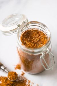 GVL An open glass jar filled with homemade taco seasoning sits on a white surface, with some of the spice spilled nearby and a measuring spoon resting beside it.