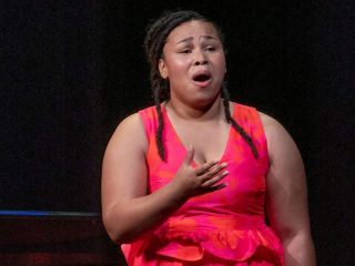 GVL A student in a bright pink dress sings passionately on stage at the Greenville Fine Arts Center, with one hand on her chest and an expressive look on her face, set against a dark background.