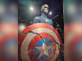 GVL A Captain America figure stands in an exhibition at the Upcountry History Museum, holding a large, cracked red, white, and blue shield with a star—just like in Avengers: Endgame. The suit is blue with a bold white star on the chest.