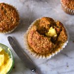 GVL A Bran Muffin with a pat of melting butter inside sits on a marble surface beside a knife, a bowl of butter, and two more Bran Muffins in the background.