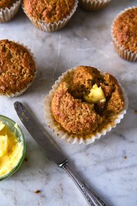 GVL A split open bran muffin with a pat of melting butter inside sits on a marble surface next to a butter knife and a small bowl of butter. More bran muffins in paper liners are visible in the background.