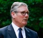 GVL A man wearing glasses, a dark suit, white shirt, and dark tie stands outside in front of green foliage, speaking or appearing to discuss topics like national security or GDP.