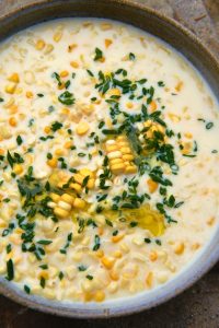 GVL A bowl of creamy corn soup, reminiscent of classic creamed corn, garnished with chopped herbs and a drizzle of oil, featuring visible corn kernels in a light, milky broth.