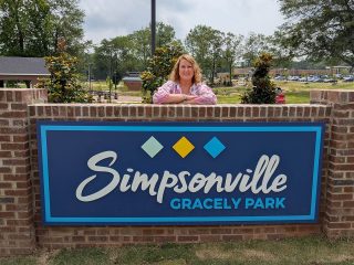 GVL A woman stands behind a brick sign that reads Simpsonville Gracely Park, honoring Dianna Gracely’s 30 years of public service, with three colorful diamond shapes above the text and trees and a park area in the background.