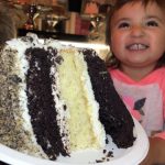 GVL A smiling young child sits behind a large slice of layered chocolate and vanilla cake with white frosting, served on a white plate—perfect for Food Baby moments or anyone craving delicious cake slices in NYC.