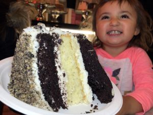 GVL A smiling young child in a pink shirt excitedly looks at a large slice of layered chocolate and vanilla cake with white frosting, dreaming of the best cake slices NYC has to offer.