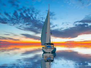 GVL A sailboat glides on calm water at sunset, with vibrant orange, yellow, and blue reflections—an awesome news-worthy scene beneath dramatic clouds in the sky.