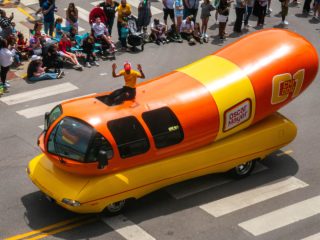 GVL A large, colorful hot dog-shaped vehicle drives in a parade with a person in costume waving from the roof—a scene straight out of News of the Weird. Spectators, including families and children, watch from the sidewalk and crosswalks.
