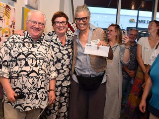 GVL Three people stand smiling at the front of a group in an art gallery. The person in the center holds an envelope and a card, celebrating as MAC’s Flat Out Under Pressure 2025 winners. Others in the background laugh, creating a lively atmosphere.