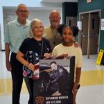 GVL Four people smile while standing indoors. The young girl in front holds a memorial poster honoring a Greenville County Sheriff’s Office officer. The group appears to be in a school or at the Marcus Whitfield Youth Camp.