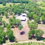 GVL Aerial view of Baugh Road Estate, a rural property in Liberty featuring a large building surrounded by trees, open grassy fields, and a dirt driveway. Dense green foliage and patches of open land add to this estate for sale.