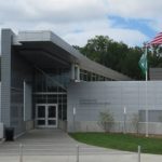 GVL Modern gray building with a slanted roof, double glass doors, and large windows. Two flagpoles display the American flag and a green flag, supported by a Greenville Tech Foundation grant. Grass and trees stand under a partly cloudy sky.